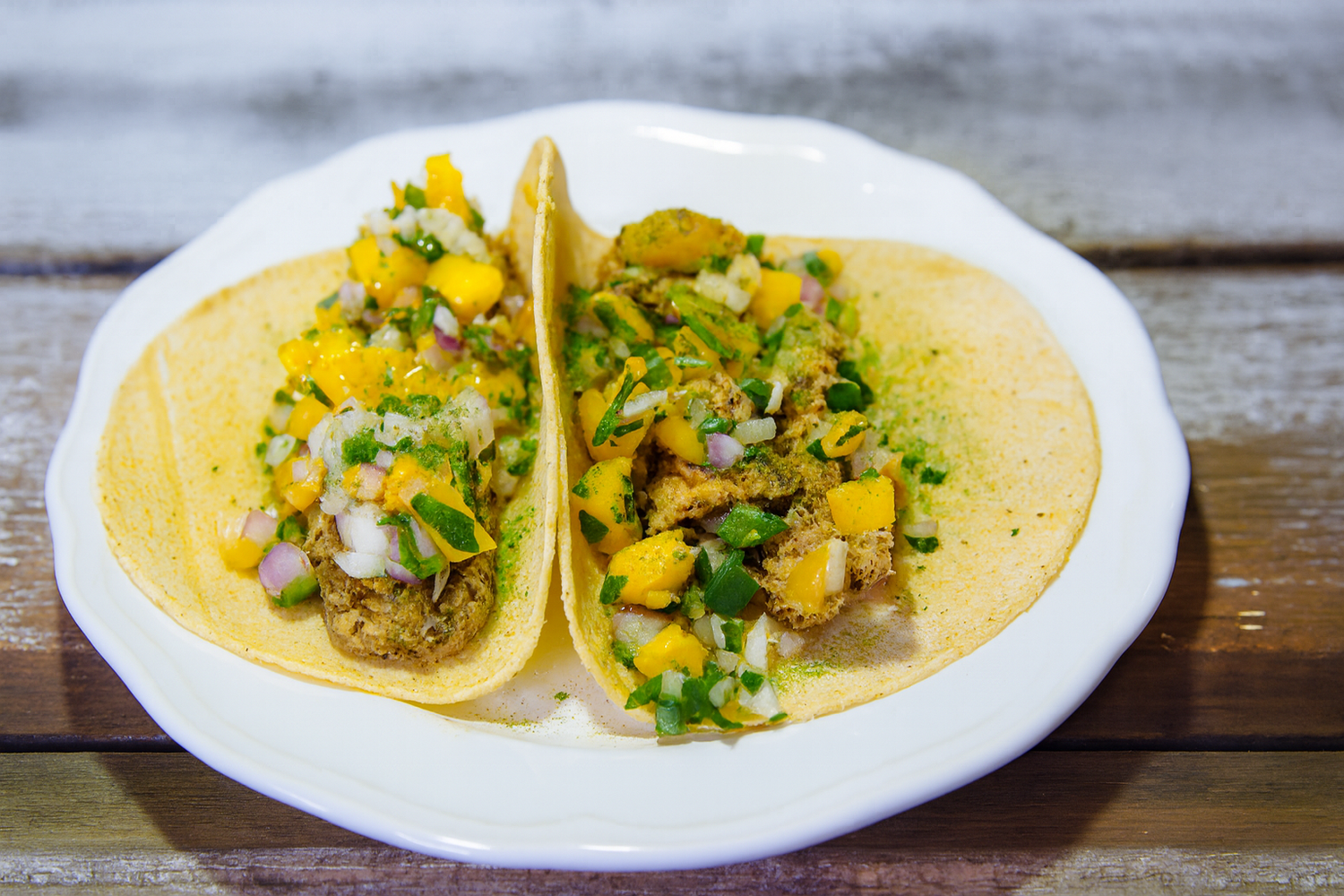Two tacos with yellow sauce and green toppings on a white plate, placed on a wooden surface.